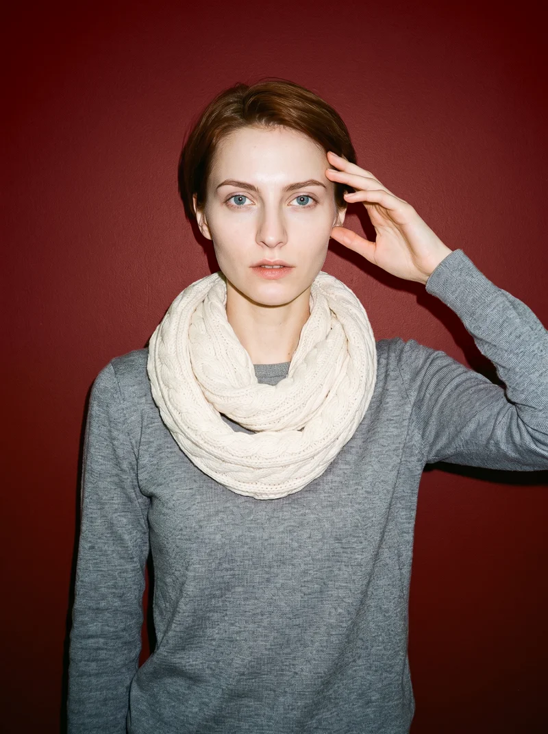 A 24-year-old Slavic woman wears a cream cable-knit snood in an upper-body, front-view composition. One hand rests at her temple, and she has a natural facial expression against an oxblood studio-wall backdrop.