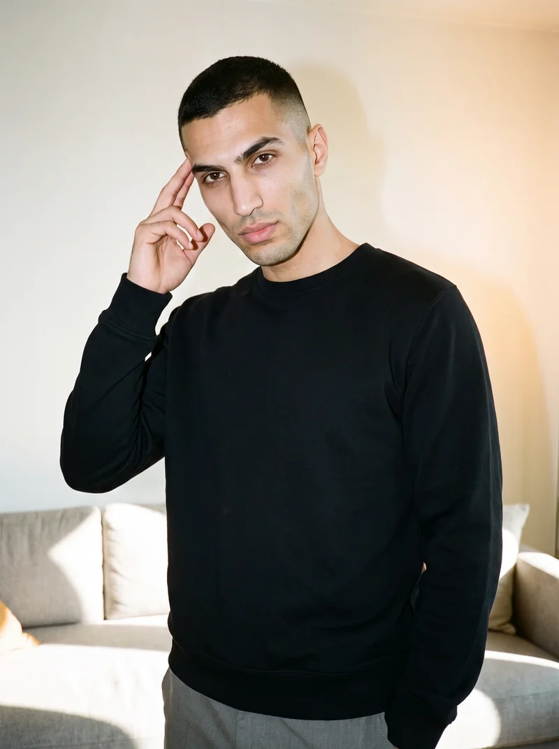 A 24-year-old Arab man is shown from the upper body at a front three-quarter angle, wearing a black crewneck sweatshirt with one hand at his temple and a confident expression. A home living room serves as the backdrop.