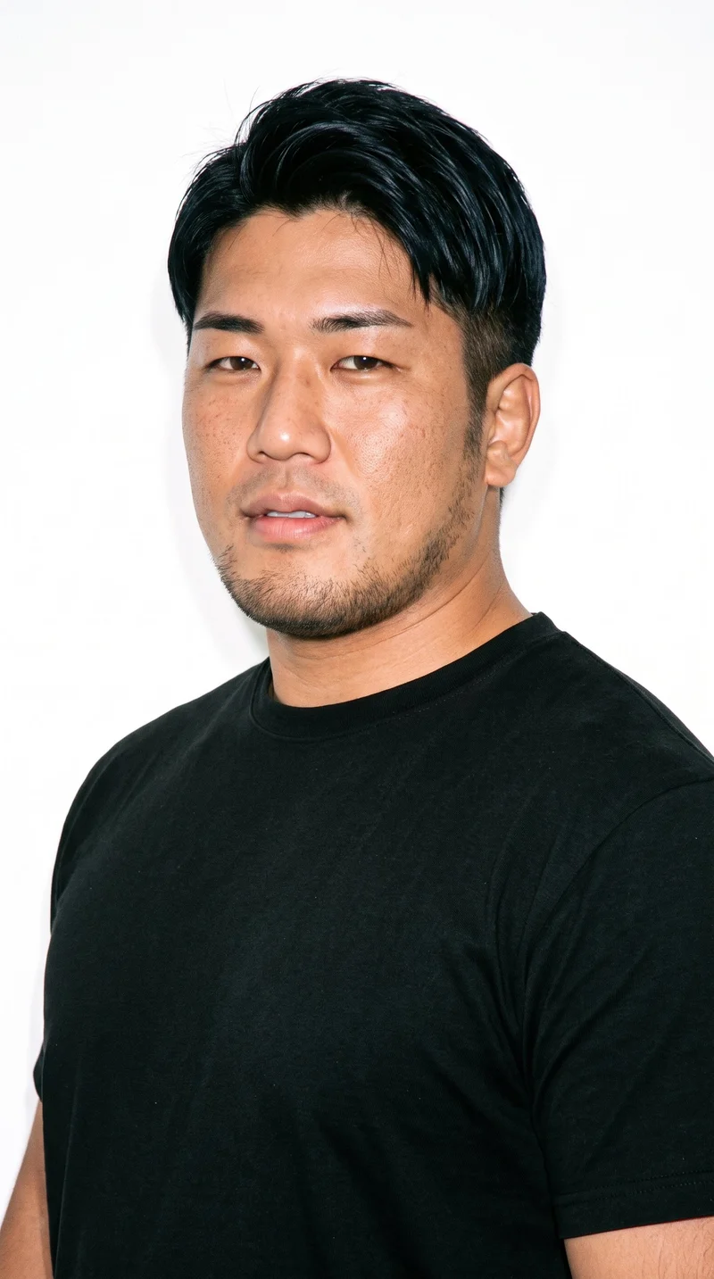 A 31-year-old Japanese man with fair skin and a neutral undertone has a rugged face with weathered, characterful features and a heavy jaw. He has thick eyebrows, very dark near-black eyes, and thin lips. His black hair is styled in a two-block cut, with short sides and back contrasted with a longer, fuller top. Very short facial-hair growth creates a faint, even shadow across the beard area.