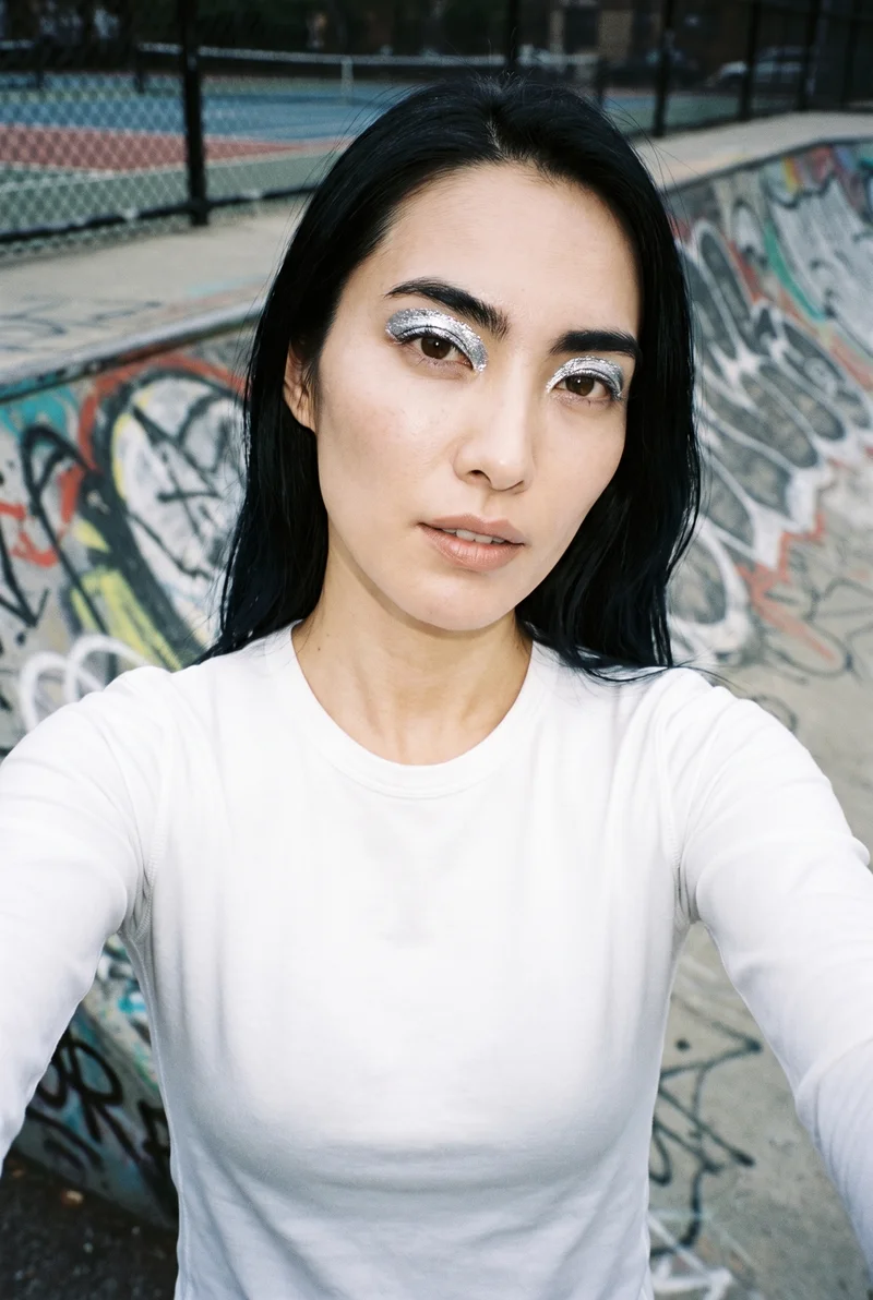 A 36-year-old Japanese woman wears a white performance base layer in a front-facing selfie. Her chin is lowered and her expression is serious, with a skate park backdrop.