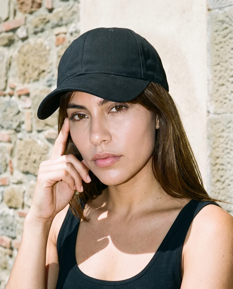 A 25-year-old Hispanic woman wears a black baseball cap, shown in a head crop from a front three-quarter angle with a finger resting at her temple and a serious expression. An Italian villa terrace serves as the backdrop.