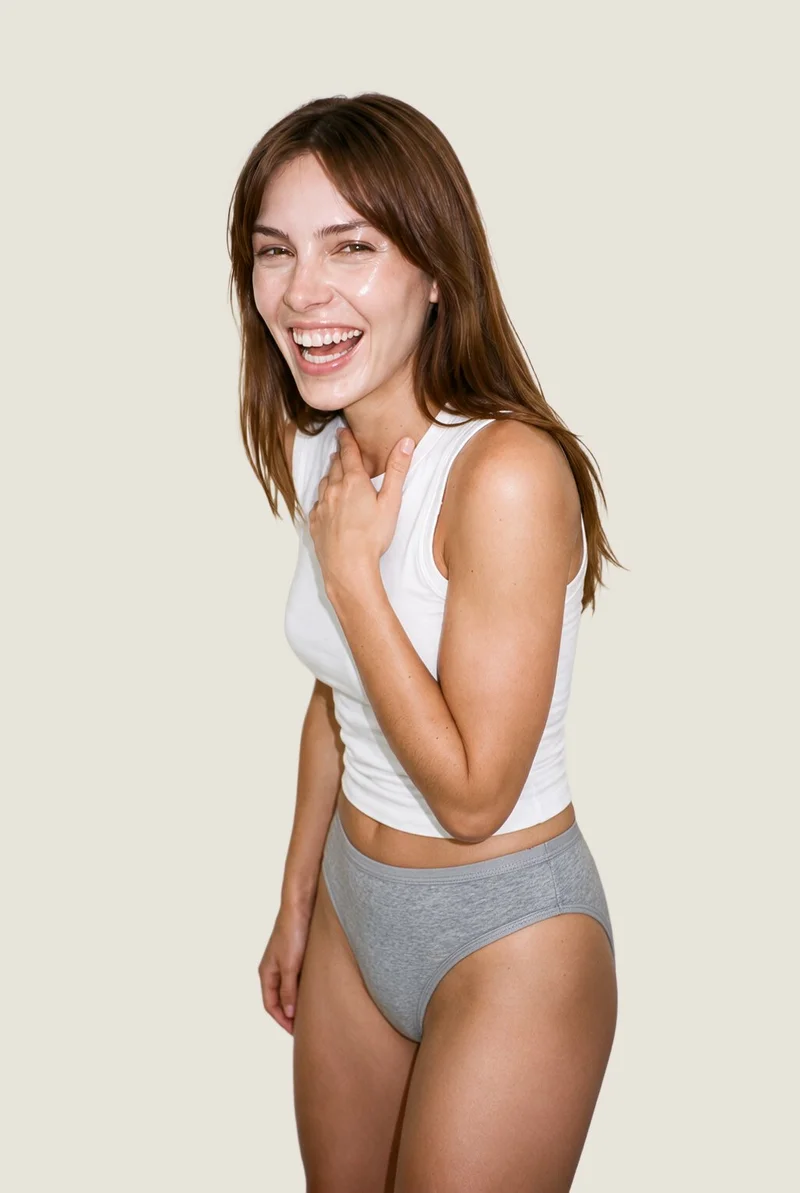 A 22-year-old Caucasian woman is shown from the upper body in an angled front view, wearing grey briefs. One hand rests at her collarbone, and she is laughing. The image has a flat Oyster Pearl backdrop.