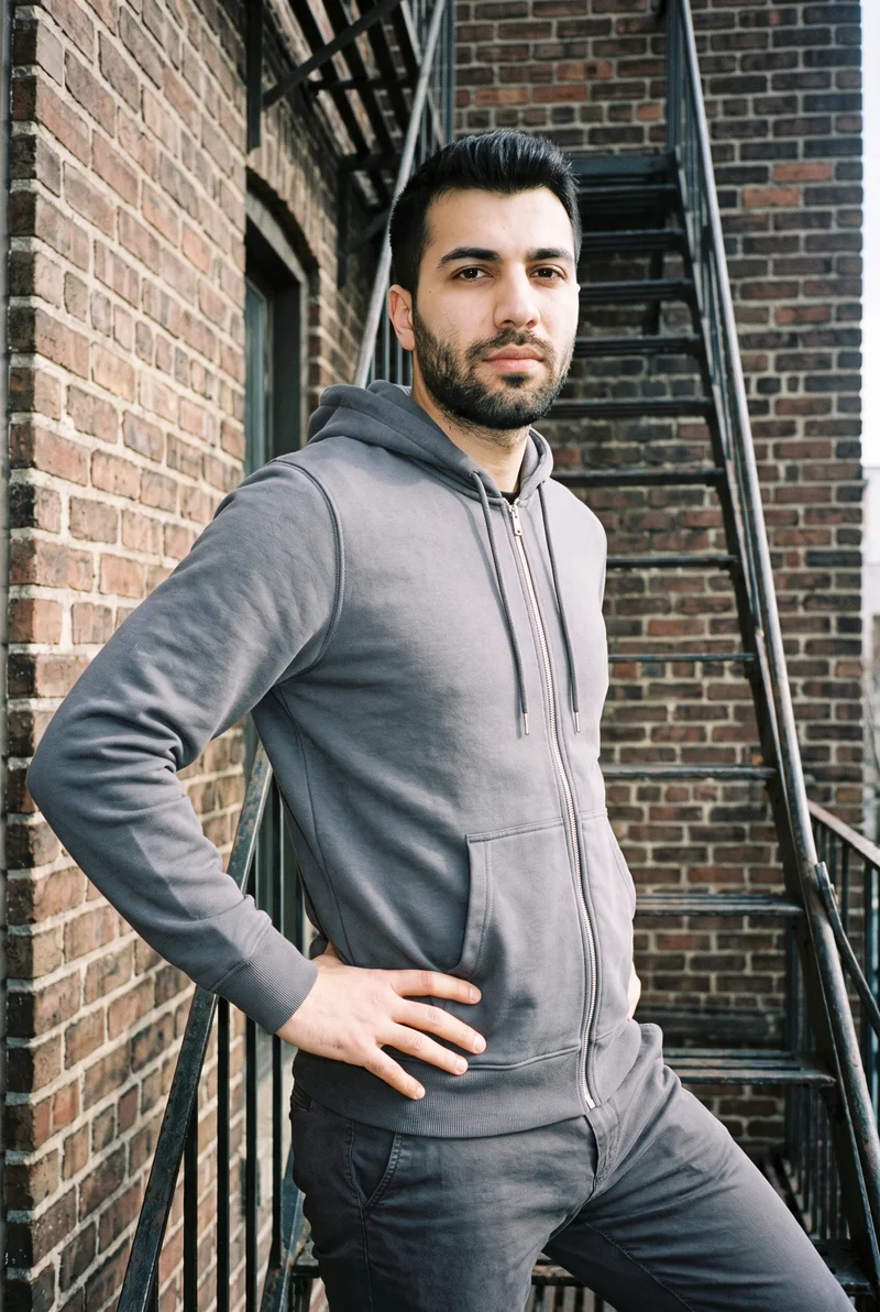 A 24-year-old Turkish man wears a charcoal cotton zip-up hoodie, shown in a three-quarter-length front three-quarter view with one hand on his waist and a neutral expression. The fire escape serves as the backdrop.