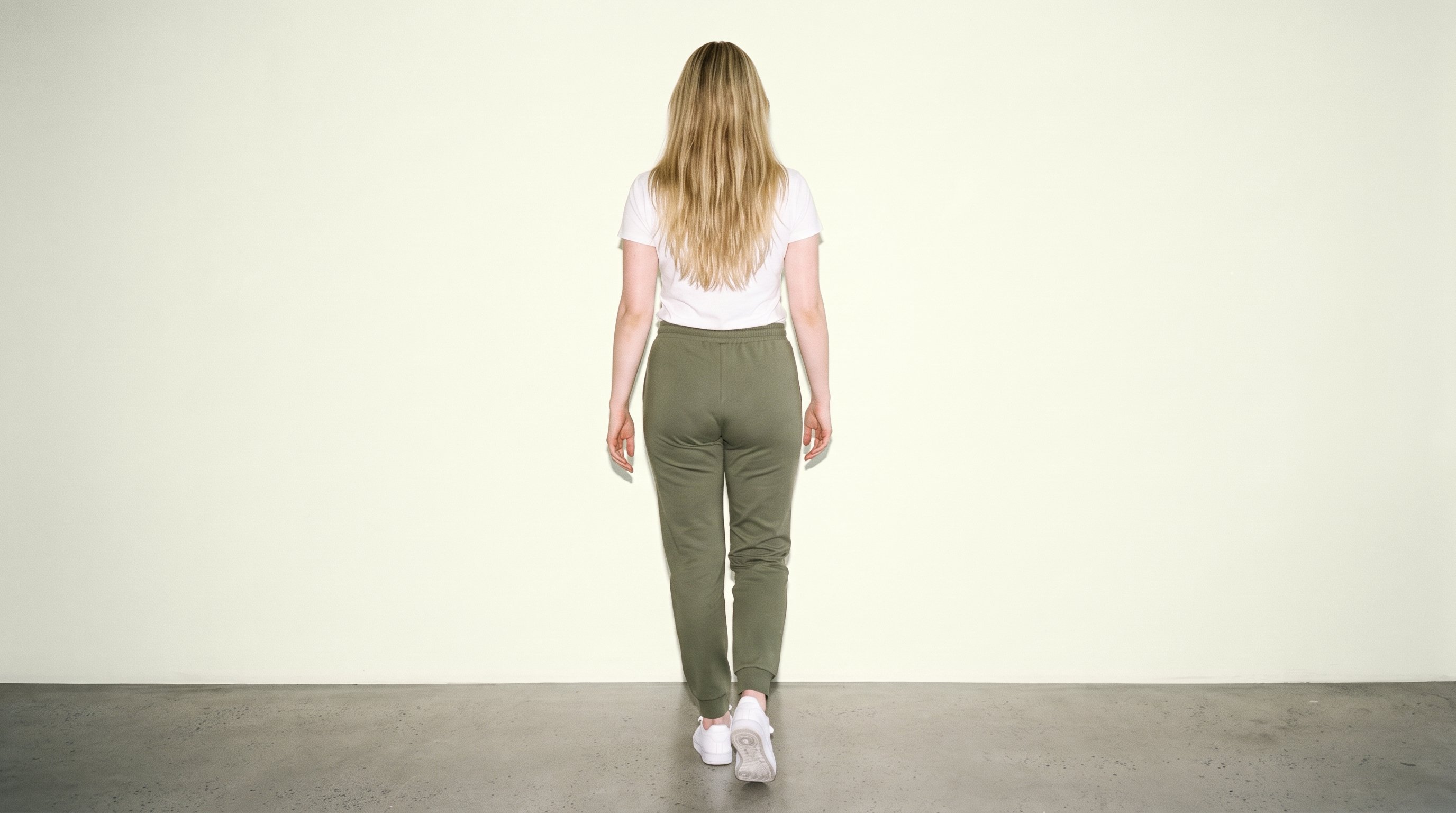 A 33-year-old Caucasian woman is shown full body from the rear while walking, wearing women's olive joggers. An ivory lace studio-wall backdrop frames the composition.