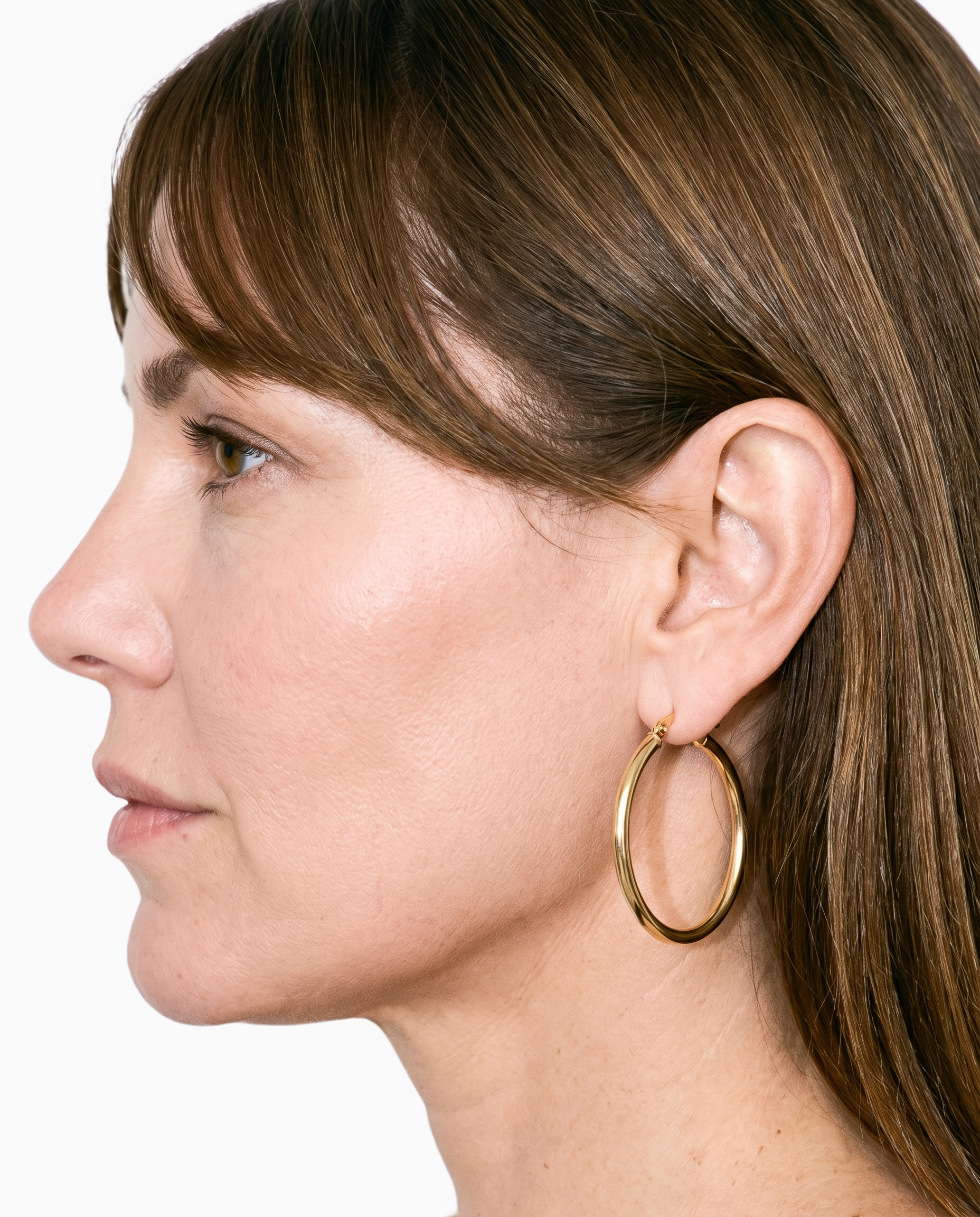 A gold hoop earring is shown on an ear from the side, with the ear positioned forward against a flat seasalt-colored backdrop.