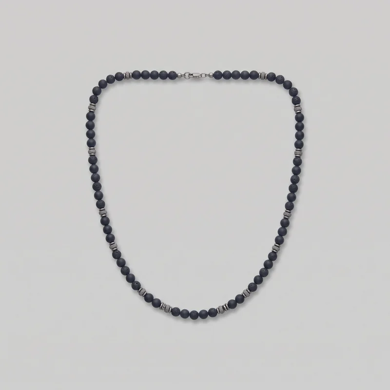 A single-strand men’s beaded necklace is laid flat in an open loop, with the clasp centered at the top. Round beads alternate between smooth and pitted textures, with repeated ribbed cylindrical and thin ring spacers arranged around the strand; smaller round beads flank the closure, and the clasp, jump rings, and connector links are visible.