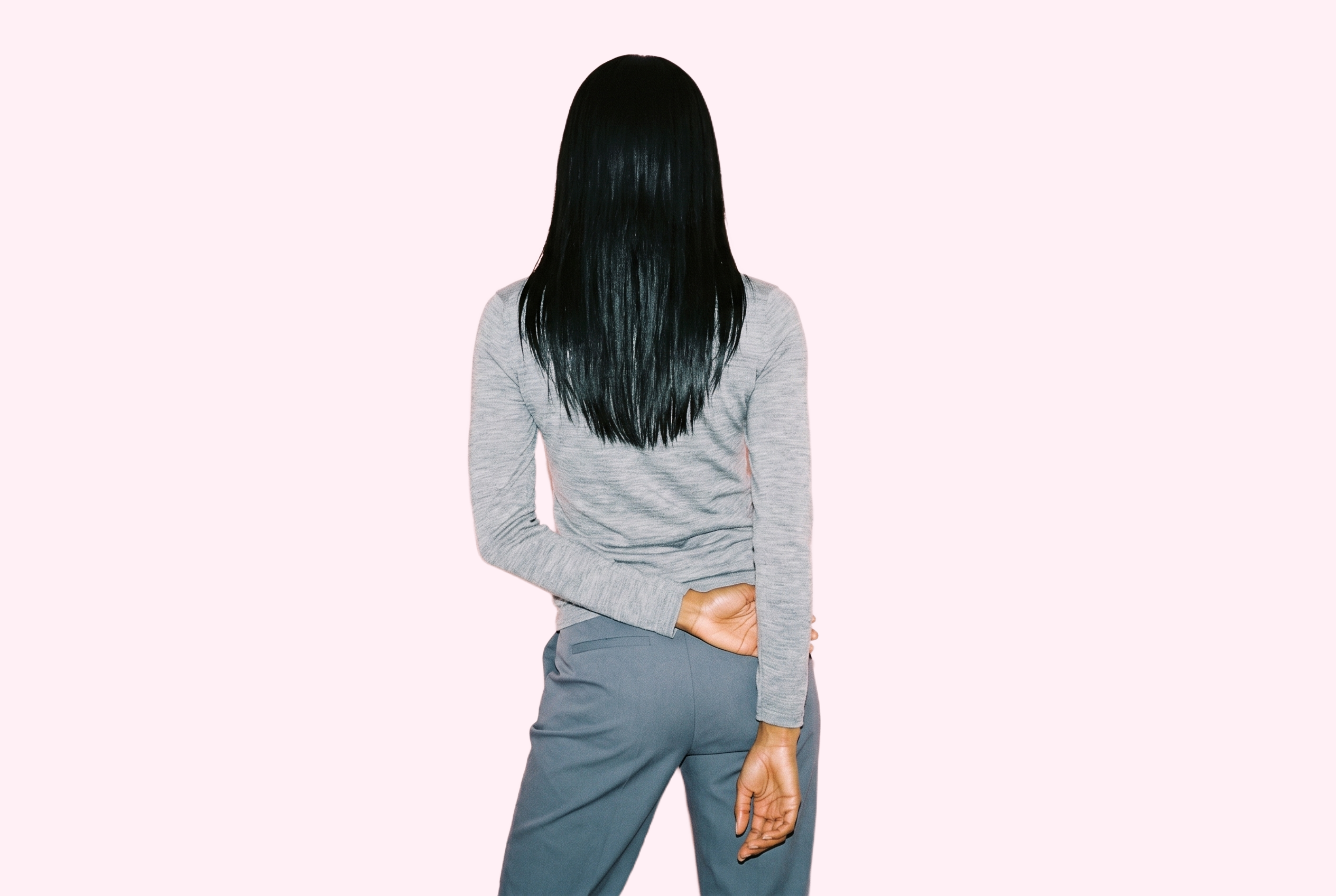 A 21-year-old African woman wears a women's grey merino thermal top in a three-quarter-length back view, standing with her hands behind her back. The lavender blush backdrop is a flat color.