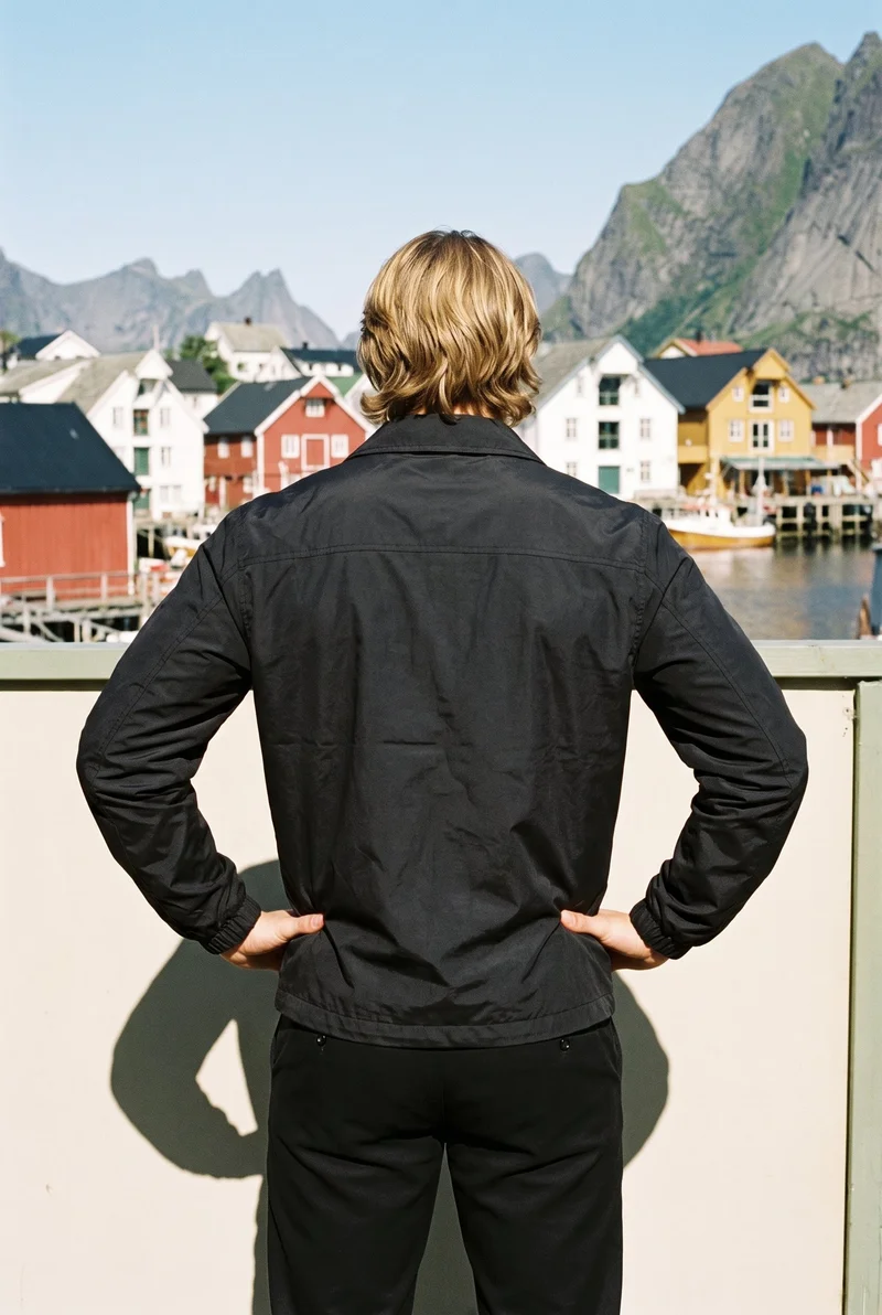 A 22-year-old Slavic male model wears a men's black windbreaker, shown from the rear in a three-quarter-length frame with his hands on his hips. A Norwegian fjord village forms the backdrop.