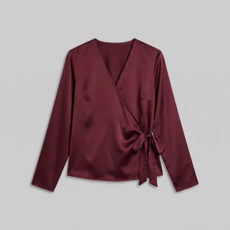 A burgundy satin wrap-front blouse is shown laid flat in a complete front view. It has a deep V neckline formed by overlapping panels, long straight sleeves, set-in shoulders, a diagonal front wrap, and an integrated side self-tie with a bow and hanging sash tails. Narrow stitched edging outlines the neckline, cuffs, and slightly curved hip-length hem, with visible bust darts or curved side seams under the arms.