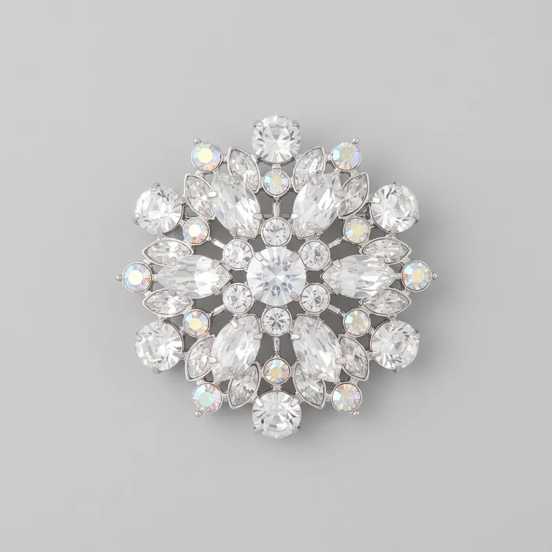 A standalone close front view shows a complete women's crystal brooch with a symmetrical radial starburst or snowflake-like silhouette. An openwork metal frame holds a central round faceted stone, surrounded by alternating round, marquise, and teardrop-shaped faceted stones in prong settings, with smaller iridescent round accents around the perimeter and inner ring. Connecting bars are visible between the stones, while the back closure is not shown.
