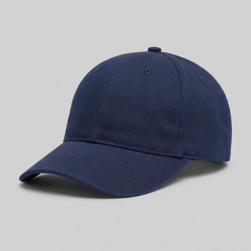 A mostly complete navy cotton baseball cap is shown in a front three-quarter view against an unspecified backdrop. The structured six-panel crown has vertical seams converging at a covered top button, with stitched ventilation eyelets on the visible panels and a raised seam along the crown base. The curved preformed brim features multiple rows of stitching, with no visible logos, prints, patches, or decorative embellishments.