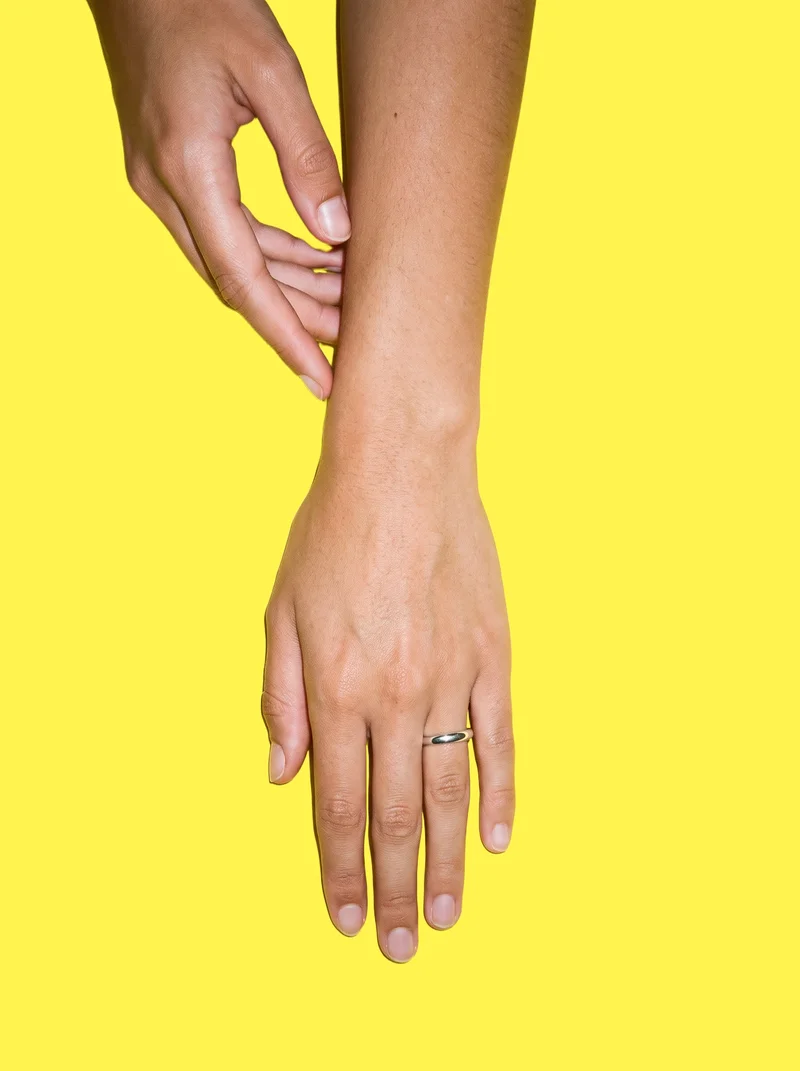 A hand and wrist area shown from a top view, wearing a women's silver band ring while touching the wrist. The subject is framed against a flat lemon-yellow backdrop.