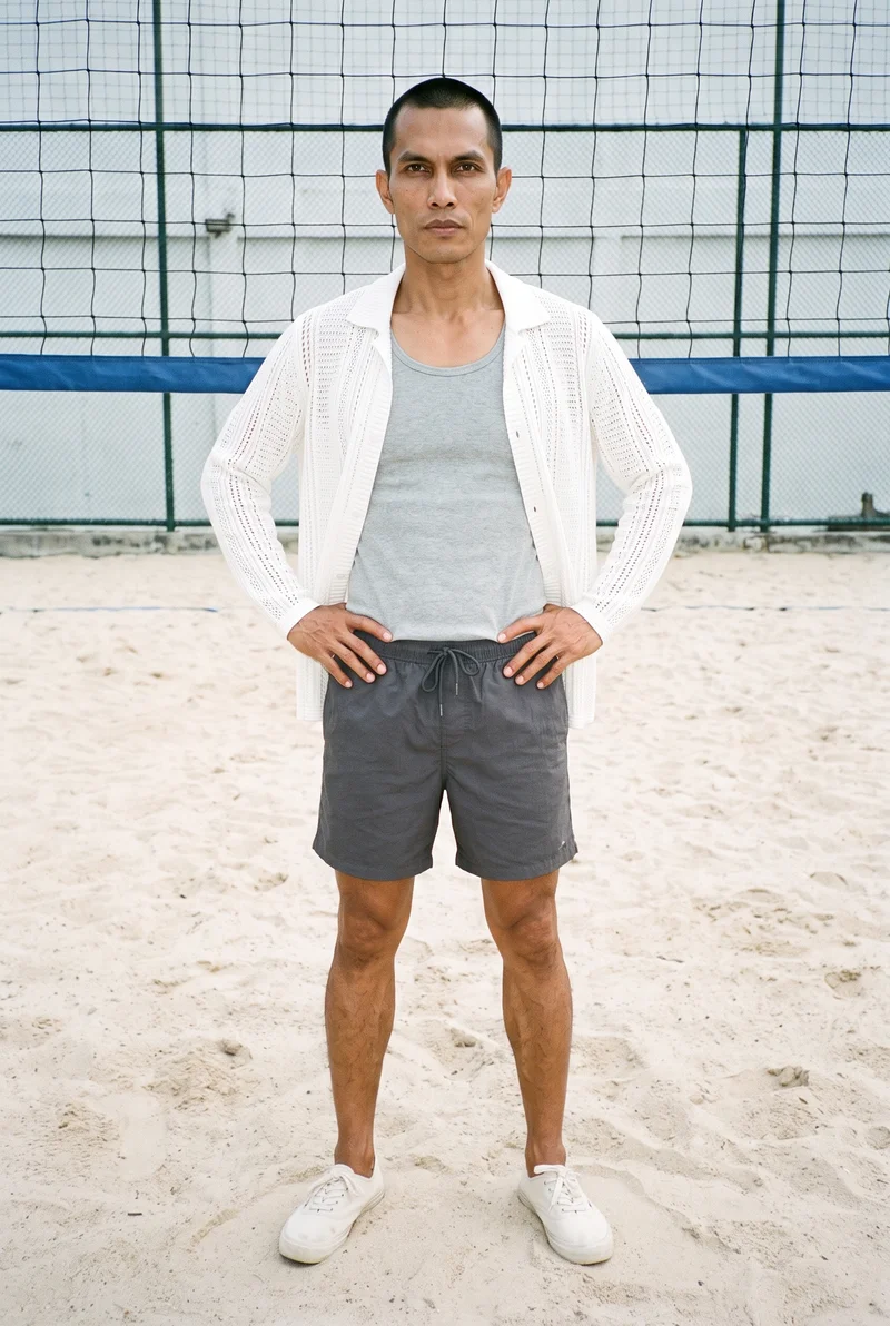 A 49-year-old Southeast Asian man wears a men’s white crochet cover-up in a full-body front view. He stands with a wide stance and an intense facial expression against a beach volleyball court backdrop.