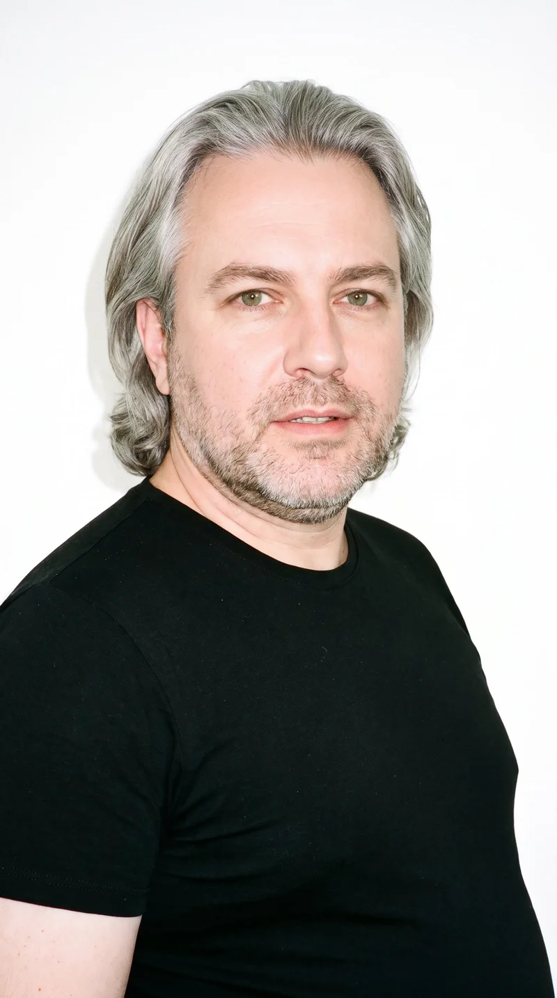A 50-year-old Caucasian man with porcelain skin and a neutral undertone has an oval face with balanced proportions and a softly rounded jaw. His medium-thickness eyebrows frame green eyes, and he has thin lips. Medium-to-long gray hair is brushed back with body and movement extending past the ears. Short, even three-day beard growth gives his facial hair a closely groomed texture.