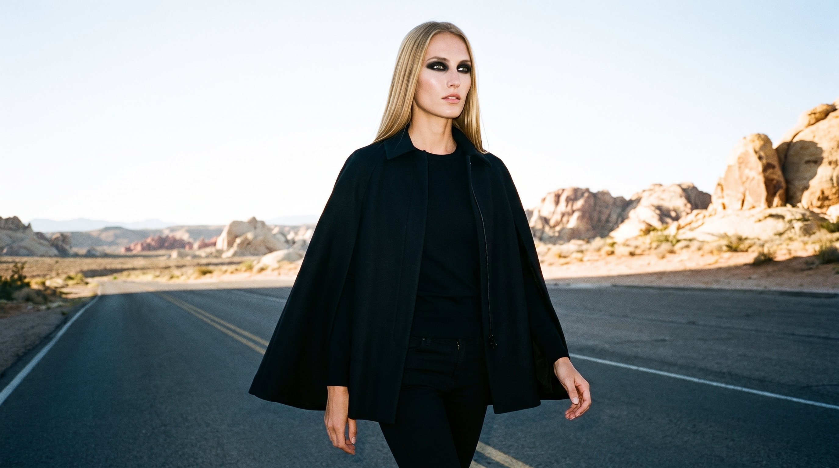 A 25-year-old Caucasian woman is shown in a three-quarter-length angled front view while walking, wearing a black cape with a natural facial expression. The Monument Valley Road backdrop frames the scene.