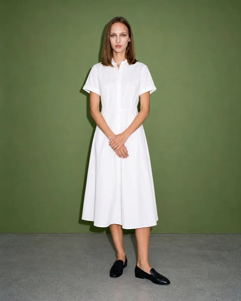 A 20-year-old Caucasian woman wears a white cotton shirt dress in a full-body front view, standing with her hands in front and a neutral expression. An olive grove backdrop is visible behind her.