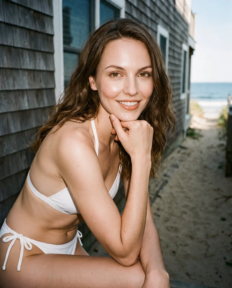 A 26-year-old Caucasian woman wears a white halter bikini in an upper-body, angled front view. She is smiling with one wrist raised to her jaw, against a Cape Cod beach house backdrop.