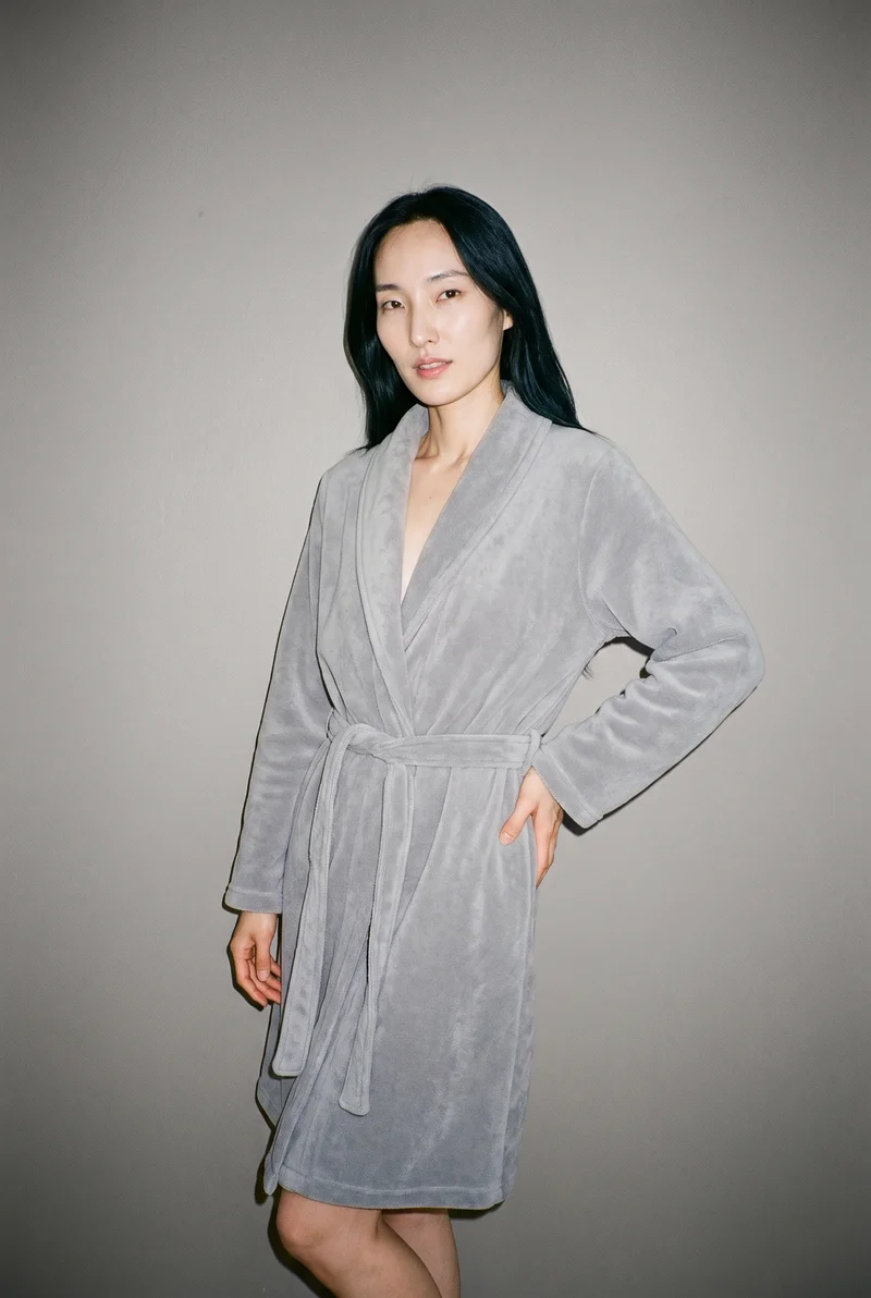 A 34-year-old Korean woman wears a women's grey fleece robe in a three-quarter-length frame, shown from a front three-quarter angle with her weight shifted back and a natural facial expression. A studio grey backdrop fills the setting.