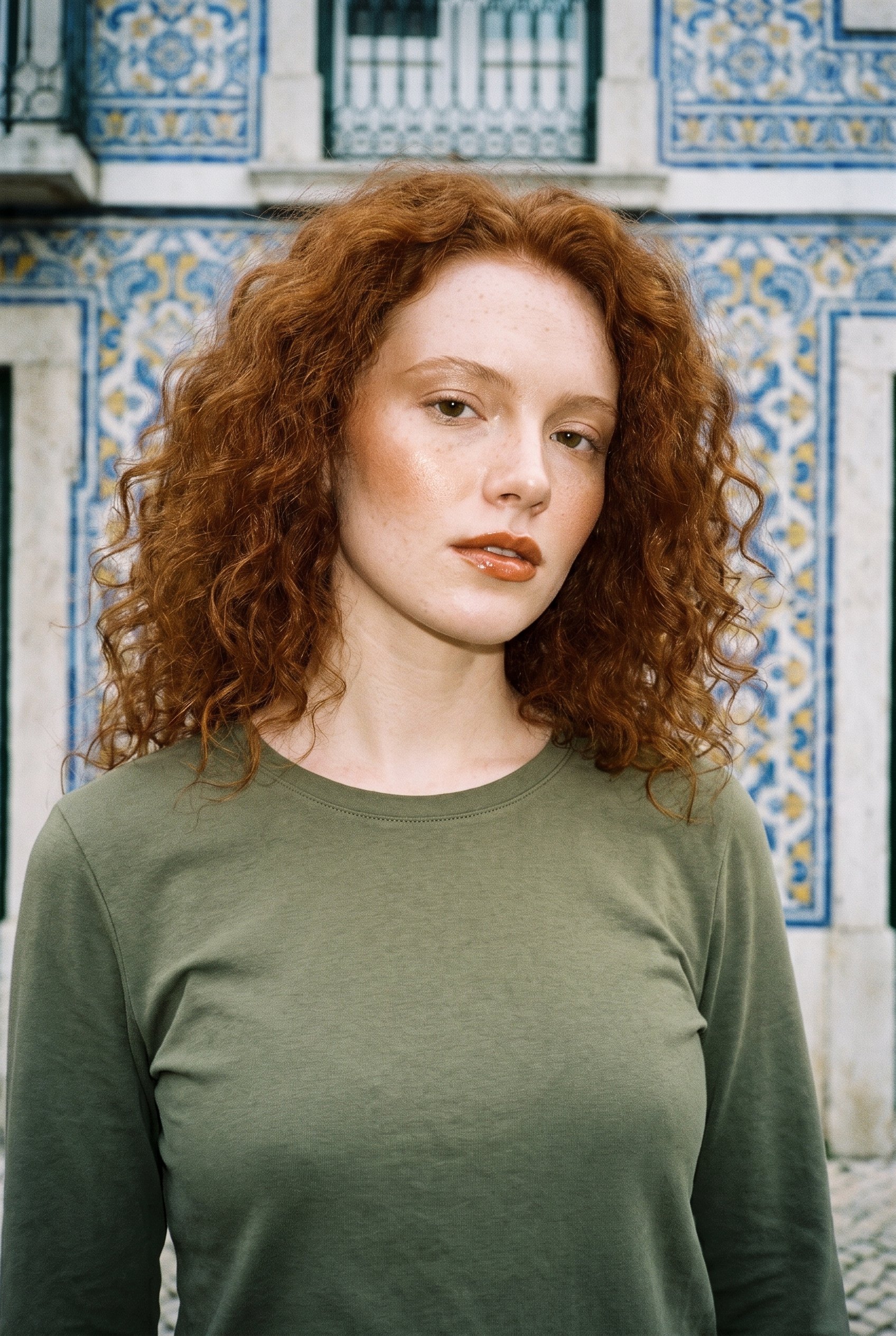 A 22-year-old Caucasian woman wears an olive cotton long-sleeve tee in a bust-framed front view. Her head is tilted with a neutral expression, against a Portuguese tile street backdrop.