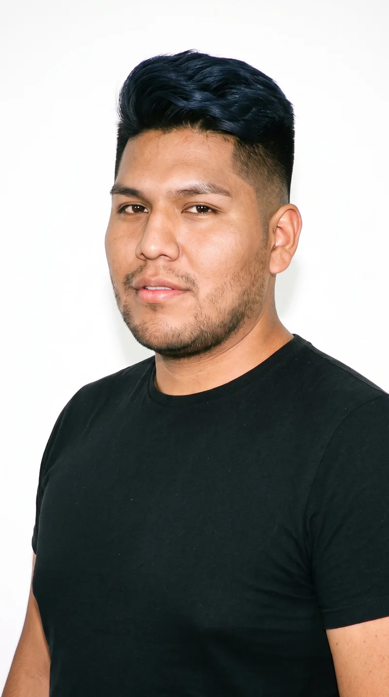 This 22-year-old Indigenous American man has brown skin with a warm golden undertone and an oval face with balanced proportions and a softly rounded jaw. His medium-thickness eyebrows frame brown eyes, and he has full lips. His black hair is styled in an undercut, with longer hair on top and markedly shorter sides and back. Dense heavy stubble forms a dark, defined layer across the beard area.