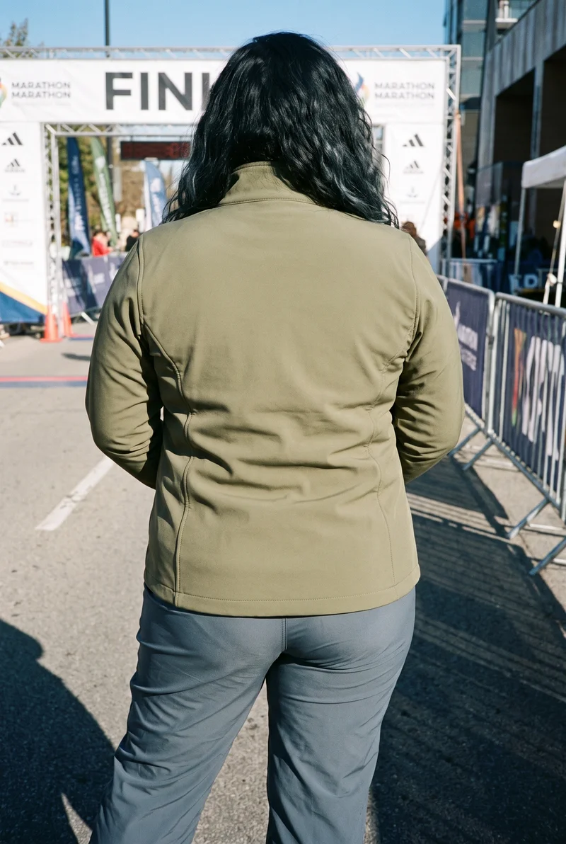 A 23-year-old Arab woman wears a women's olive softshell jacket, shown from the back in a three-quarter-length frame with her hands behind her back. A Marathon Finish Line backdrop is visible.