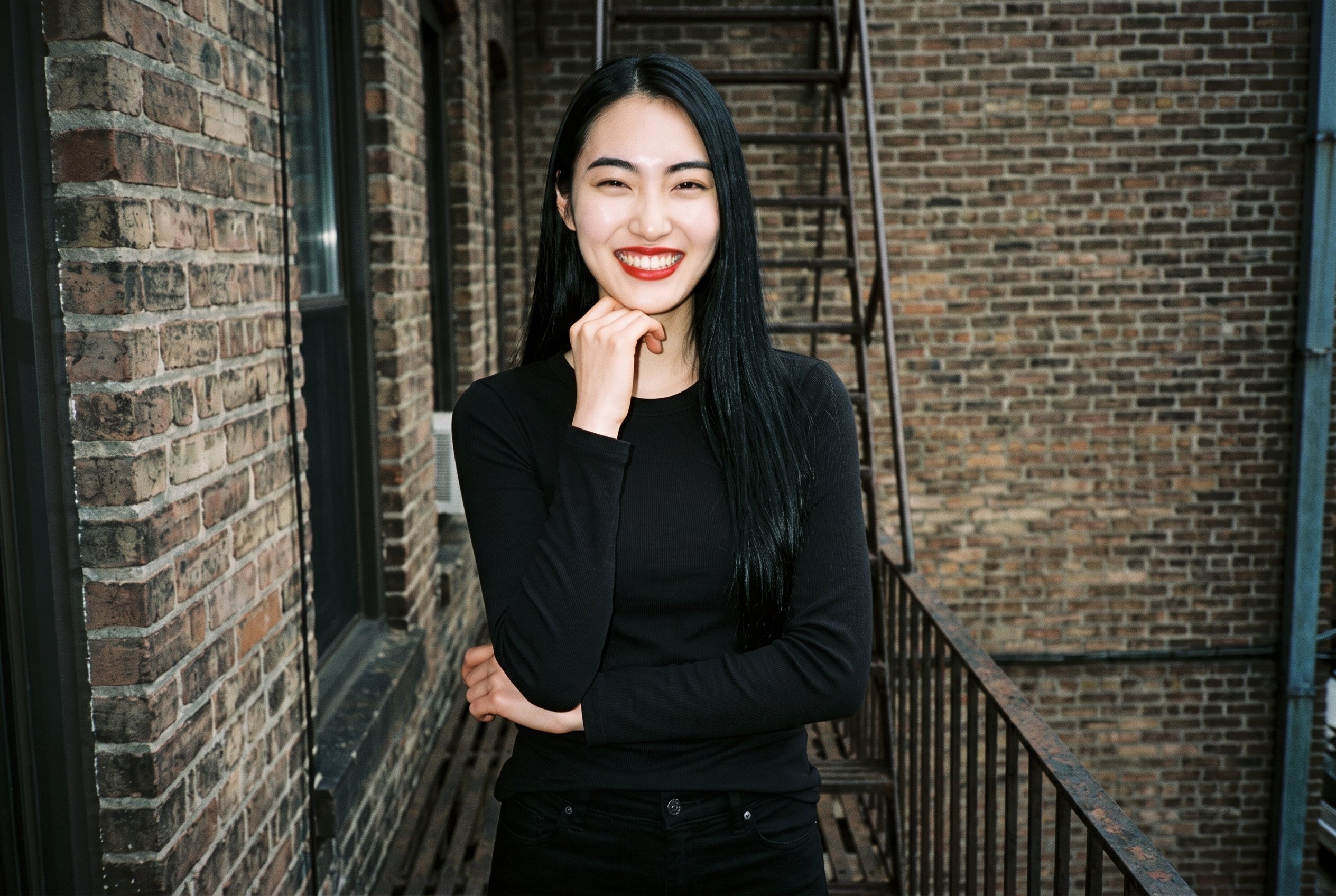 A 22-year-old Japanese woman wears a black thermal top in an upper-body, front-view composition. She grins with one wrist raised at her jaw, against a fire escape backdrop.
