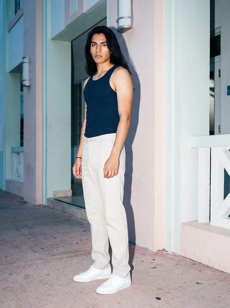 An 18-year-old Hispanic male model wears a men's navy blue tank top in a full-body front three-quarter view, standing with his ankles crossed and a neutral expression. A Miami Beach Deco Hotel serves as the backdrop.