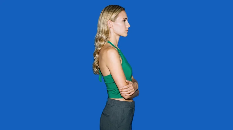 A 25-year-old Caucasian woman wears an emerald green halter top in a side-view upper-body composition. She poses with a self-hug and a natural facial expression against a flat denim blue backdrop.
