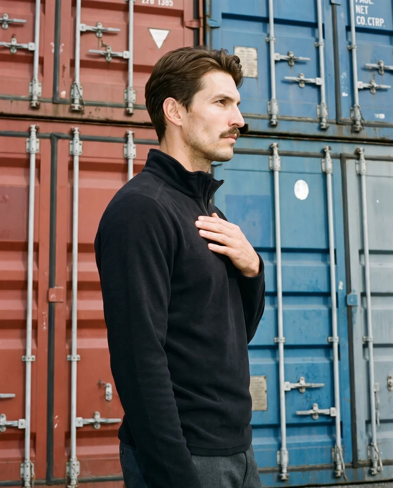 A 28-year-old Caucasian man wears a black half-zip in an upper-body side view, with his fingers at his chest and a neutral expression. Shipping containers form the backdrop.