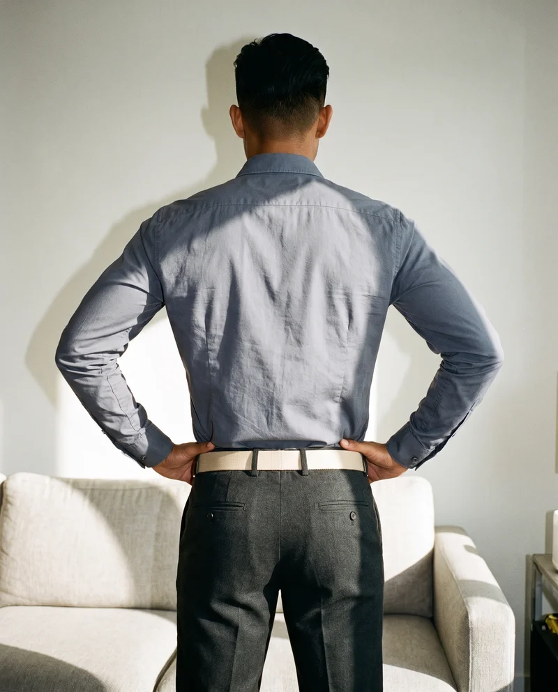 A 20-year-old Southeast Asian man is shown from the back in a three-quarter-length view, standing with his hands on his waist and wearing a beige canvas belt. A home living room serves as the backdrop.