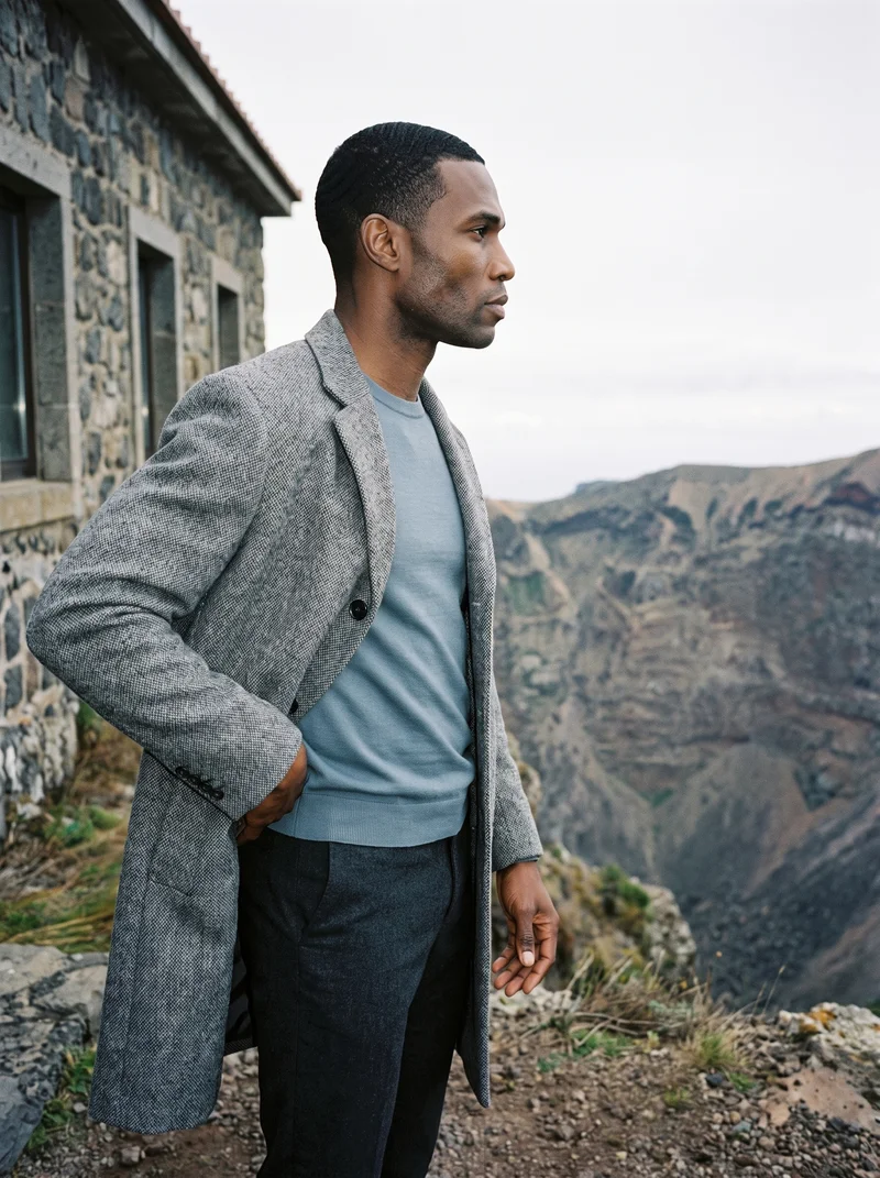 A 42-year-old African man wears a grey wool coat in a three-quarter-length side view, with one hand resting on his lower back and an intense facial expression. A Santorini caldera view forms the backdrop.