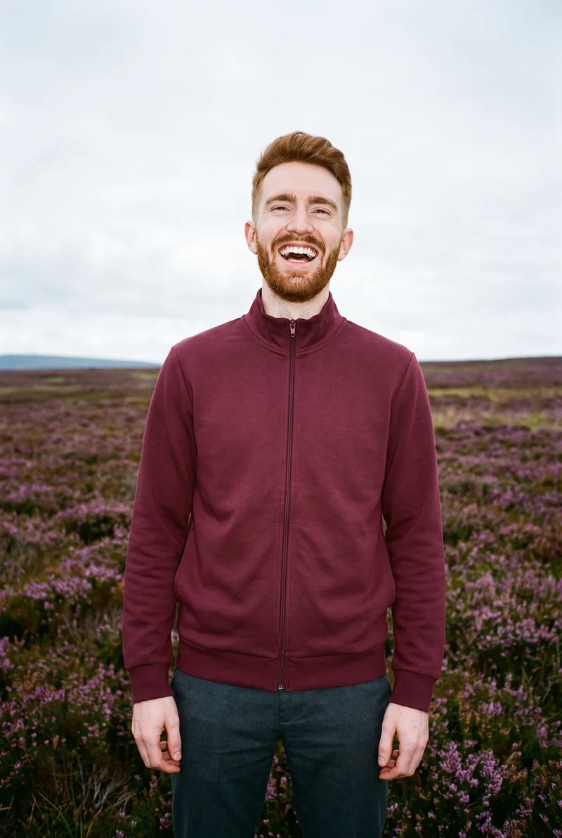 A 22-year-old Caucasian man wears a burgundy tracksuit top in a relaxed standing pose, shown from the front in a three-quarter-length frame. He is laughing, with a Scottish Highlands moor serving as the backdrop.