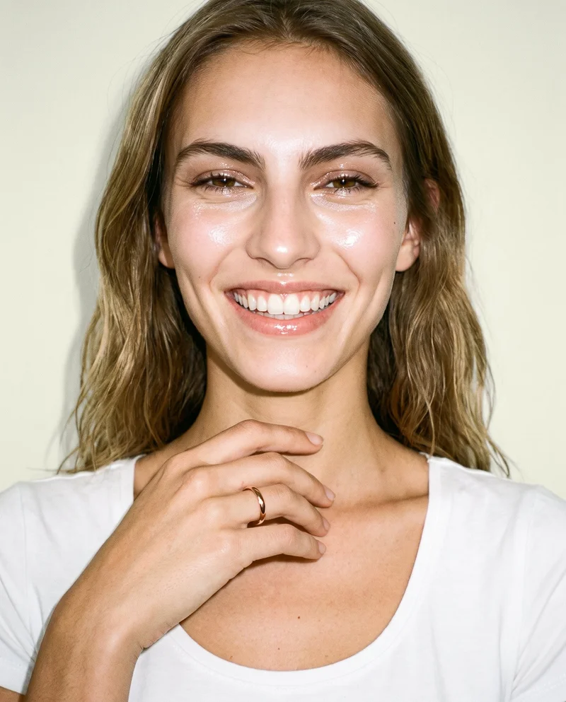A 19-year-old Caucasian woman is shown in a front-facing bust view with one hand raised to her collarbone, displaying a women's rose gold band. She has a grin, and an ivory lace backdrop fills the background.