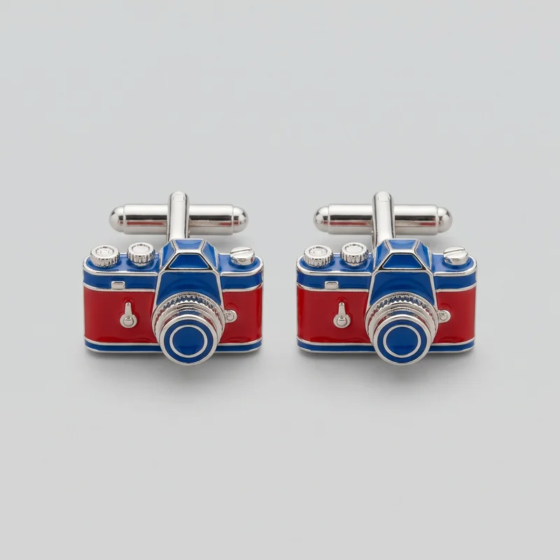 A matching pair of camera-shaped enamel cufflinks is shown from the front, with their T-bar swivel backs visible behind the camera faces. Each cufflink has a rectangular camera body, glossy enamel panels, polished metal edging and outlines, raised circular lenses with concentric rings, top control knobs, and a raised viewfinder-like center section with small front relief details.