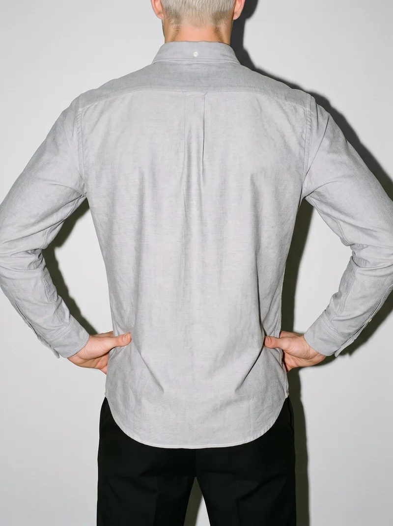 A male model is shown from the rear in a torso-framed view wearing a grey cotton Oxford shirt, with his hands on his hips. A pearl grey studio-wall backdrop fills the background.