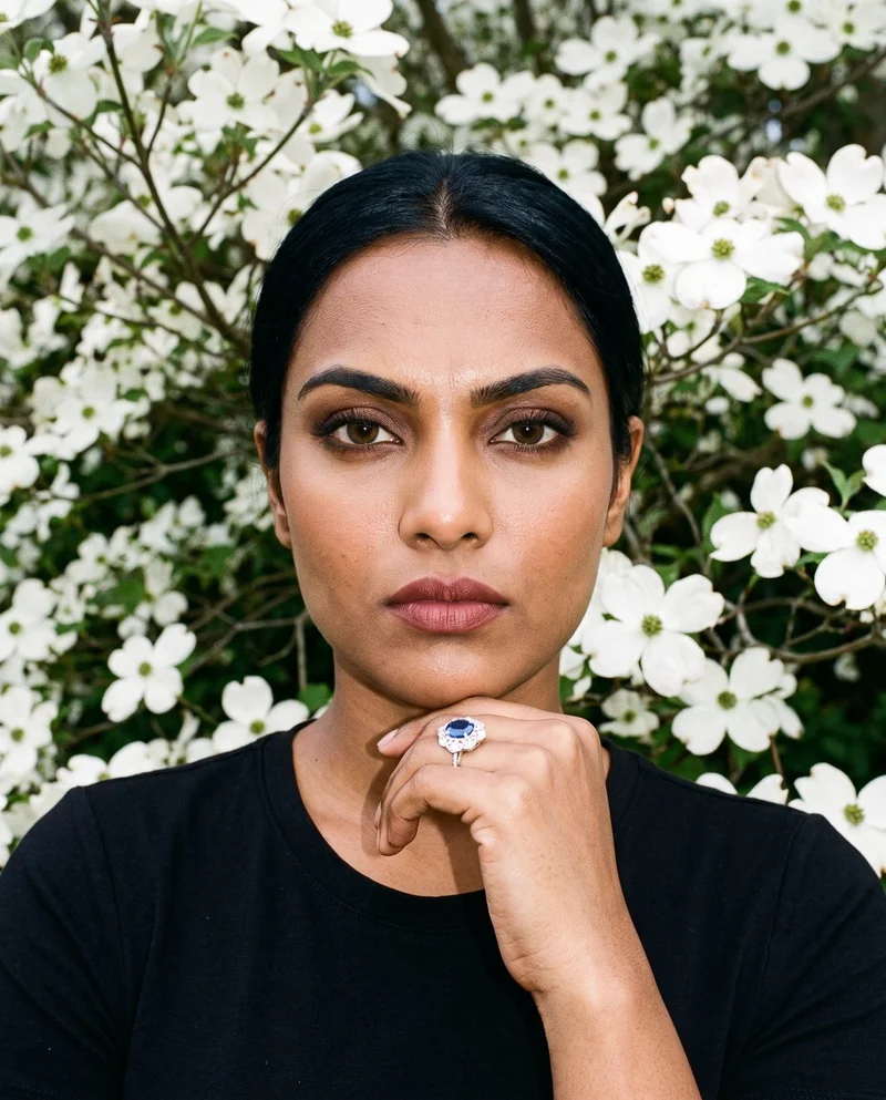 A 28-year-old Indian woman is shown in a front-view bust composition with her wrist raised beside her jaw, displaying a gemstone statement ring. Her expression is intense, with a dogwood tree grove forming the backdrop.