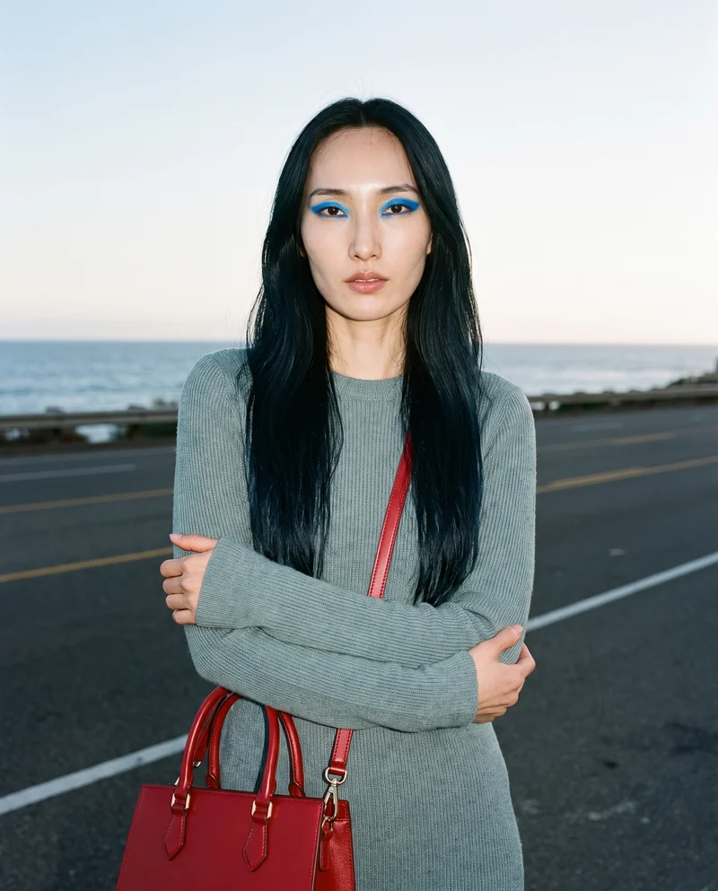 A 34-year-old Korean woman is shown from the upper body in front view, holding a red top-handle handbag at her elbow. Her confident expression and the Pacific Coast Highway backdrop are visible.