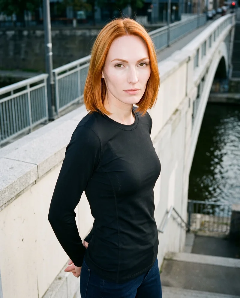 A 25-year-old Caucasian woman wears a black compression base layer, shown from an angled front view in an upper-body composition. Her hands are behind her back and her expression is natural. A pedestrian bridge backdrop is visible.