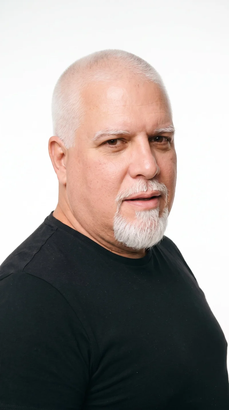 A 56-year-old Caucasian man with medium skin and a warm golden undertone. His face has a chiseled, angular structure with a defined jaw and pronounced cheekbones. He has thick eyebrows, brown eyes, and full lips. His white hair is clipped into a very short, even buzz cut, and he has a compact goatee centered on the chin with clear cheeks.