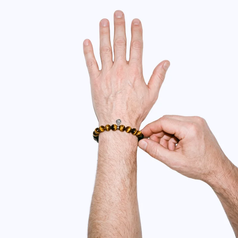 A man's hand and wrist are shown from above while he touches the men's gemstone beaded bracelet worn around his wrist. The bracelet and wrist area are framed against a flat porcelain backdrop.