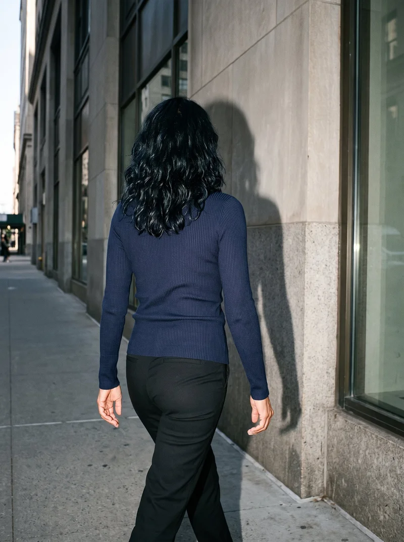 A 45-year-old Hispanic woman wearing a women's navy ribbed turtleneck is shown from the rear in a three-quarter-length frame while walking. The Financial District urban canyon forms the backdrop.