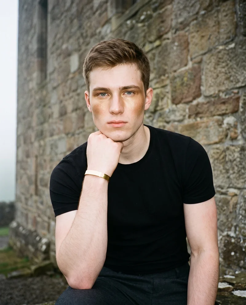A 19-year-old Caucasian man is shown in an upper-body front view with a neutral expression, holding his wrist beside his jaw to display a men's wide gold cuff. A Scottish castle serves as the backdrop.