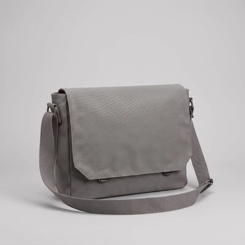 A grey canvas messenger bag is shown standing alone from a slight front angle. Its structured rectangular body has a boxy satchel shape, a flat base, straight side panels, and a large fold-over flap with angled lower corners, edge binding, and visible border stitching. Two small rectangular clasp tabs appear beneath the flap, while the shoulder strap is draped around the bag with ring attachments and an adjustable slider visible.
