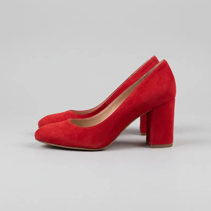 A pair of red women's closed-toe court pumps is shown in a side three-quarter view. The shoes have rounded almond toes, low-cut openings with open insteps, closed backs, high straight block heels, slim sole edges, and fine stitching around the toplines.