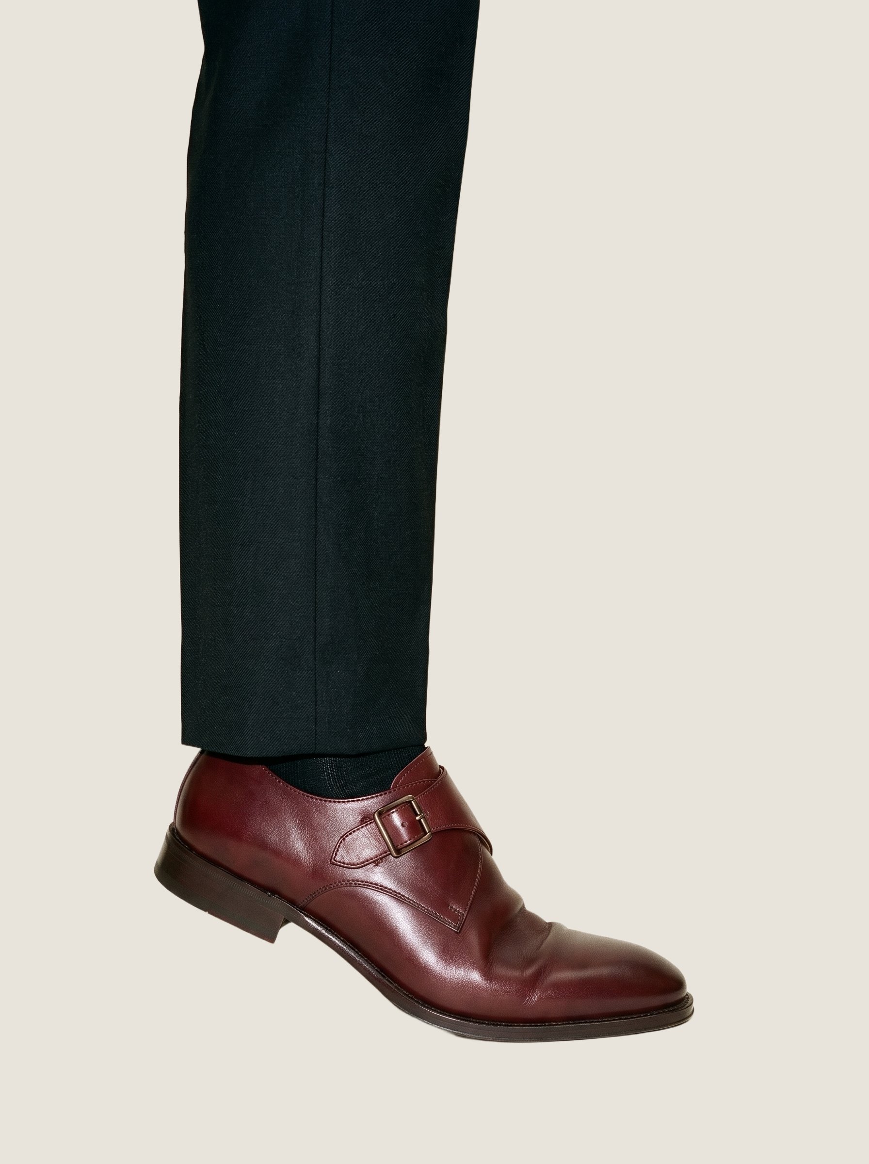 Oxblood monk-strap shoes shown in an ankle-framed side view while positioned on tiptoes, against an oyster pearl flat-color backdrop.