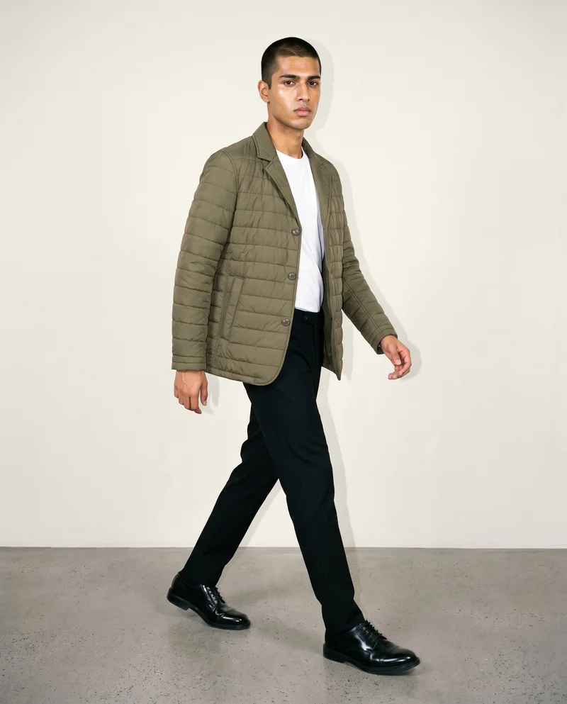 An 18-year-old Indian man wears an olive quilted jacket in a full-body angled-front view while walking with a serious expression. The jacket is shown against an Oyster Pearl studio-wall backdrop.
