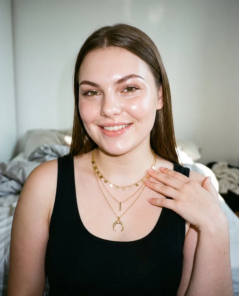 A 19-year-old Caucasian woman is shown in a front-facing bust view, smiling with one hand at her collarbone to display a women's gold layered necklace. A bedroom backdrop frames the composition.