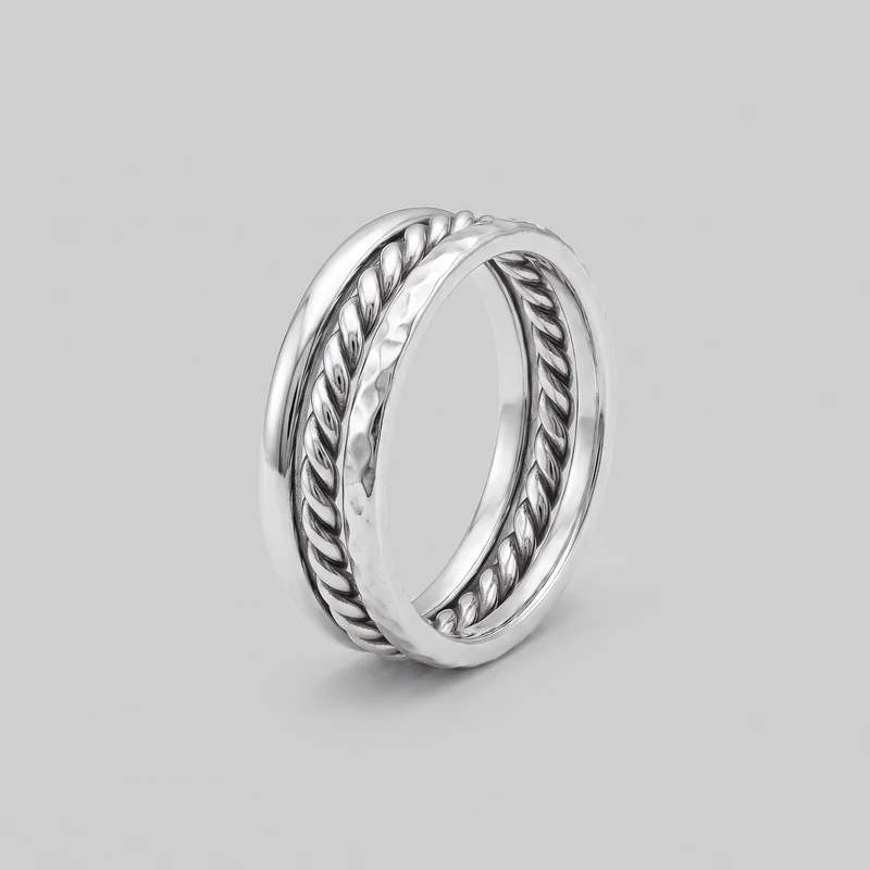 A complete silver stacking ring is shown upright at an angle, with its circular opening and rounded profile visible. Concentric raised rails and recessed channels surround the band, including a continuous braided-link motif bordered by smooth rounded bands and an uneven hammered-texture raised band. No stones, logo, print, engraving, or closure are visible.