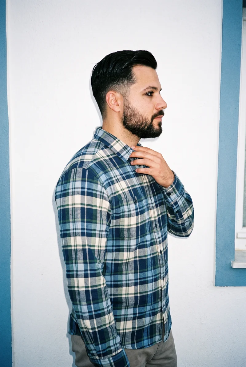 A 26-year-old Caucasian man is shown in an upper-body side view wearing a blue tartan flannel shirt, with one hand resting at his collarbone. His natural expression is visible against a Greek Villa White Blue backdrop.