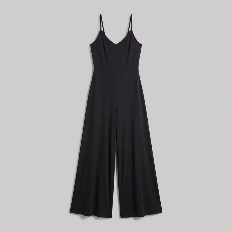 A black sleeveless wide-leg jumpsuit is shown in a full front standalone view against a plain backdrop. It has a fitted bodice with a deep V neckline, narrow shoulder straps, low armholes, a horizontal waist seam, diagonal bust and side seams, and a center front seam extending through the full-length pants. The pant legs hang away from the body and end in wide, straight hems.